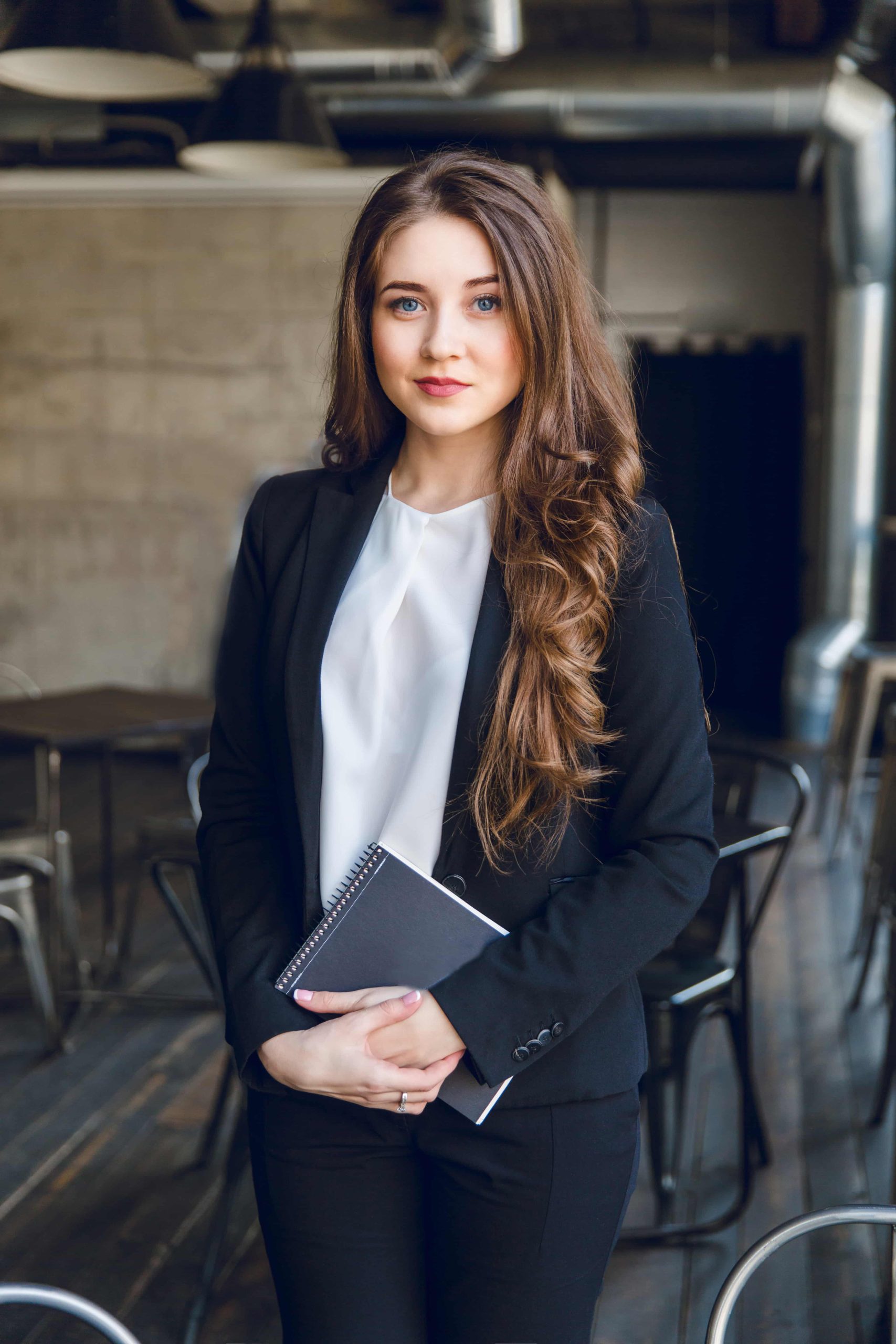 brunette-business-woman-with-wavy-long-hair-blue-eyes-stands-holding-notebook-hands-min